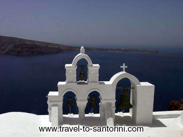 OIA CHURCH BELLS - The view of Roka on Thirassia from behind a church in Oia. Genuine sample of the cycladic architecture we find on churches. by Ioannis Matrozos